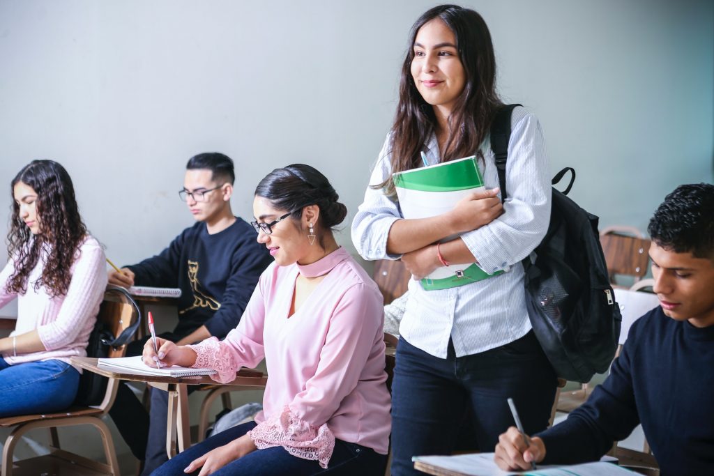 Students sitting at desks, one standing up with a white and green textbook