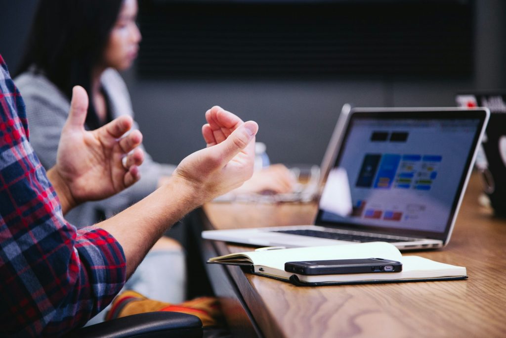 Photograph of hands waving in a meeting conversation with a computer screen and phone in the foreground