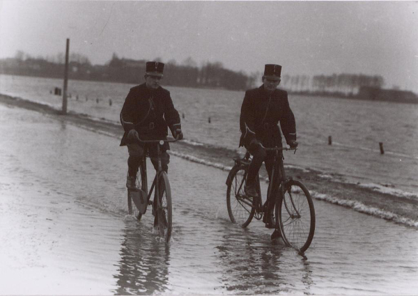 Two people crossing of the flooded Elftweg in 1930 on bikes, in black and white.