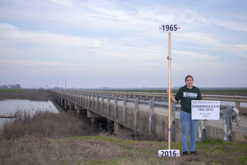 Photograph of a person in front of a bridge holding a pole indicating how much the land has dropped between 1965 and 2016